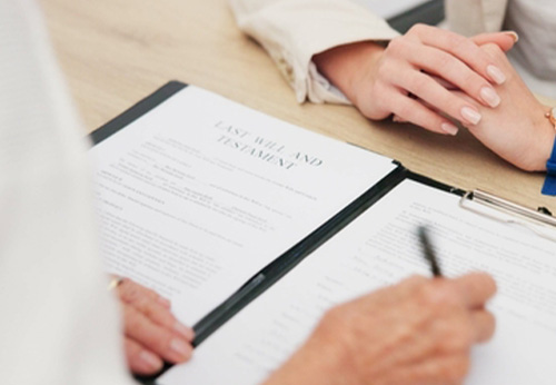 A younger man helps an older man review and sign a document at a table.